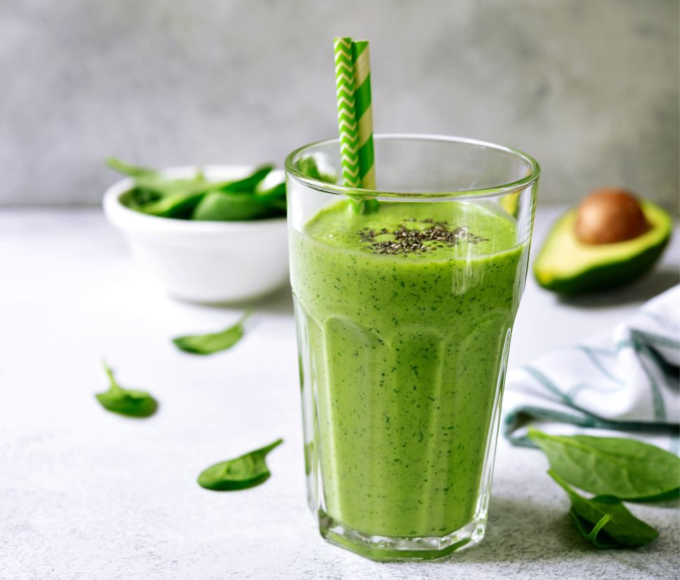 A close-up of a fertility-friendly IVF green smoothie for hormone balance, served in a tall glass with two green paper straws. The smoothie is vibrant green, topped with a sprinkle of chia seeds. In the background, there’s a halved avocado, fresh spinach leaves, and a white bowl filled with more spinach, all resting on a light grey surface with a soft, striped cloth nearby.