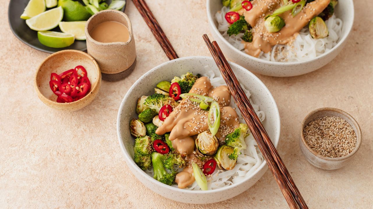 A fertility-friendly cashew ginger chicken dish served in a speckled ceramic bowl over white rice noodles, topped with roasted Brussels sprouts, broccoli, sliced red chilli, spring onions, and sesame seeds. A pair of wooden chopsticks rests on the bowl. Surrounding the dish are small bowls of sesame seeds, sliced red chilli, and a creamy cashew sauce, along with a plate of fresh lime wedges and spring onions. This nourishing meal is packed with protein, healthy fats, and fertility-boosting nutrients to support reproductive health.