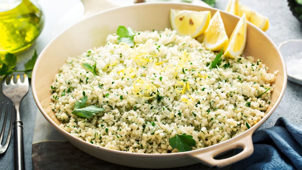 A beige ceramic pan filled with lemon herb cauliflower rice, garnished with fresh parsley and lemon zest. The bowl is placed on a rustic grey table with a fork, olive oil, and lemon wedges in the background, creating a fresh Mediterranean-inspired setting.