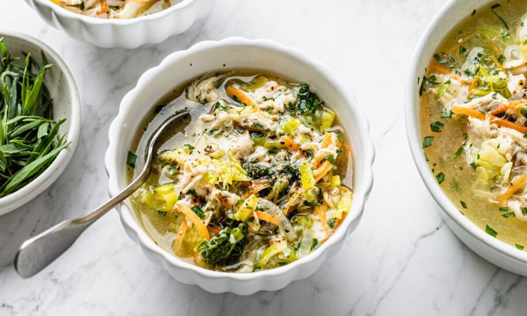 A bowl of homemade bone broth with shredded chicken, carrots, cabbage, and fresh herbs in a white scalloped bowl. A spoon rests inside the bowl, ready for a comforting sip. Another bowl of the same nourishing soup and a dish of fresh green herbs are visible in the background on a white marble surface.