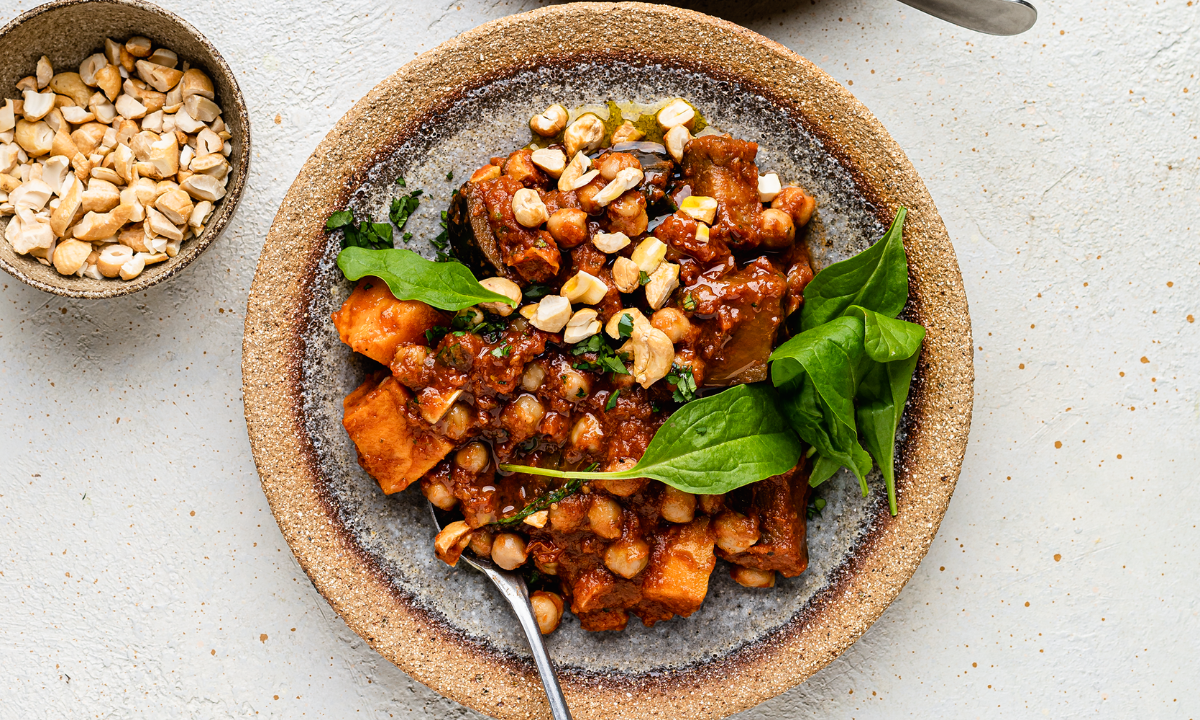 A nourishing bowl of chickpea and aubergine stew with sweet potatoes, garnished with fresh spinach leaves and chopped cashews. The dish is served in a rustic ceramic bowl, with a side of crushed cashews in a small bowl on a textured, neutral-toned background. The rich, spiced tomato-based sauce glistens, highlighting the depth of flavour in this wholesome, fertility-friendly meal.