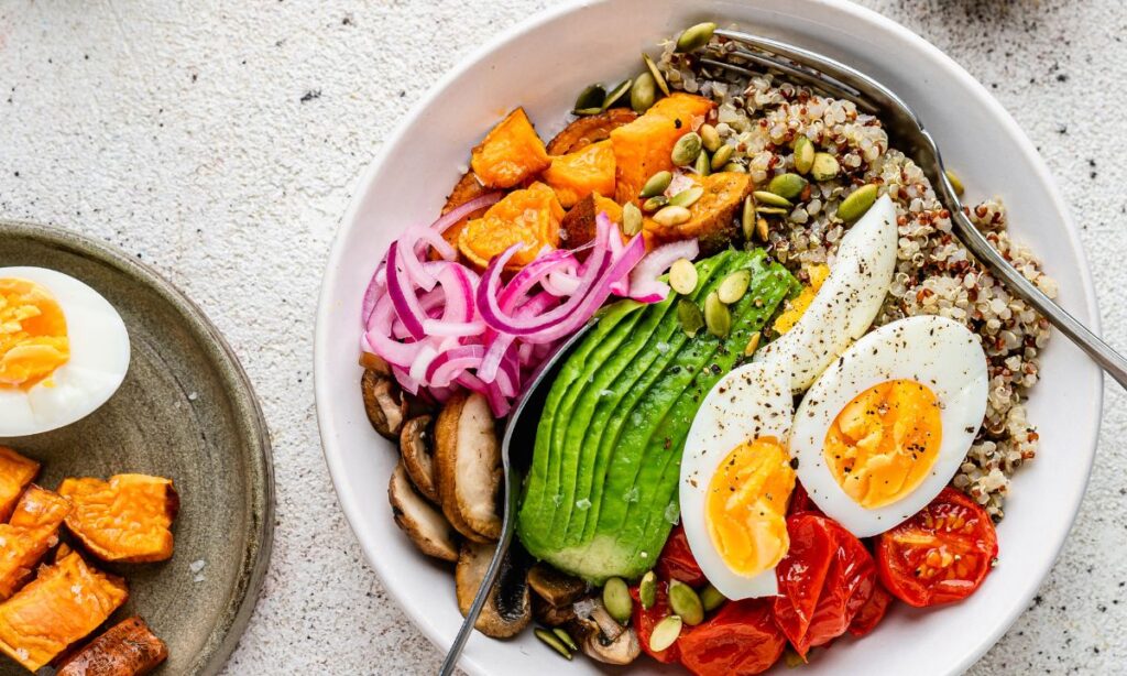 A vibrant bowl featuring quinoa topped with avocado slices, roasted sweet potato cubes, sautéed mushrooms, cherry tomatoes, pickled red onions, pumpkin seeds, and a halved soft-boiled egg. The bowl is arranged artfully, with a fork resting on the side, alongside a smaller plate of additional roasted sweet potato cubes and a halved boiled egg. The dish is presented on a light, textured surface, showcasing a nutritious and colourful meal.