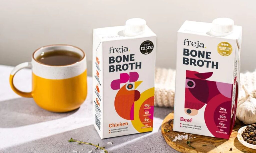 Alt text: "Two cartons of Freja Bone Broth, one chicken and one beef, placed on a light surface with a yellow mug filled with bone broth. The packaging features modern, minimalist illustrations of a chicken and a cow. A wooden board with garlic, peppercorns, and sea salt is nearby, with a cozy knit blanket in the background."