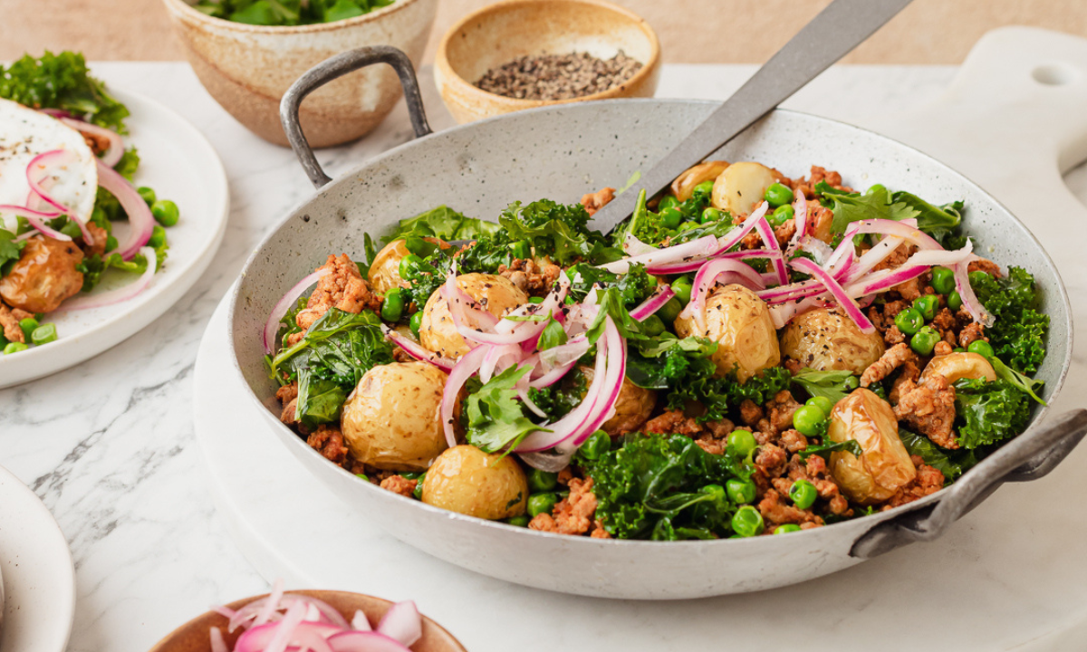 A nourishing iron-rich breakfast hash served in a rustic pan, featuring golden roasted baby potatoes, sautéed kale, peas, spiced minced meat, and topped with thinly sliced red onions and fresh herbs. A plate in the background showcases a serving of the hash with a fried egg on top. A wholesome, protein-packed meal designed to support fertility and boost iron intake naturally.