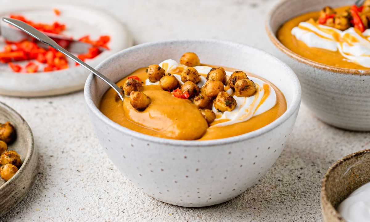 A creamy orange-hued sweet potato soup served in a speckled ceramic bowl, topped with crispy spiced chickpeas, a dollop of yoghurt, and a drizzle of sauce. A spoon is dipped into the soup, highlighting its thick texture. In the background, additional bowls of soup and a plate of chopped red bell pepper are visible, set on a neutral-toned surface.