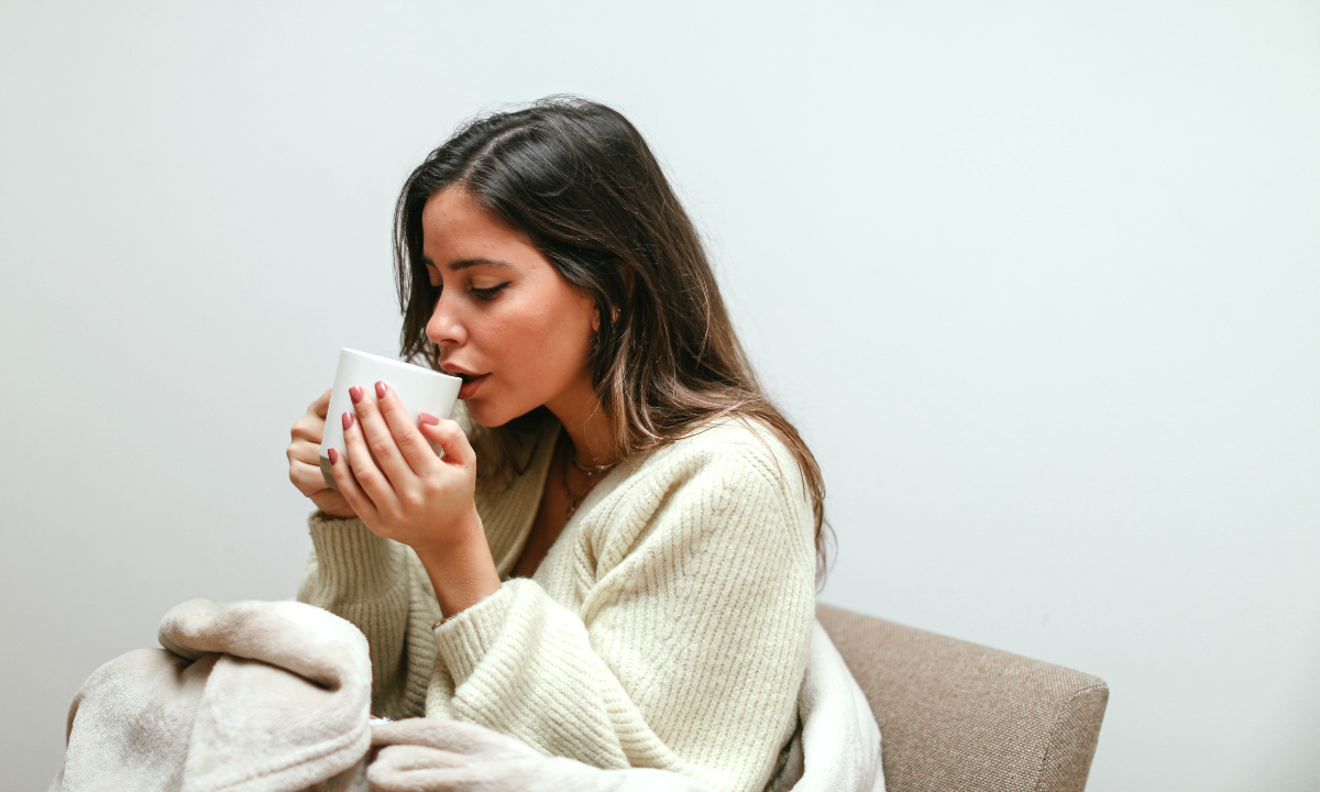 A young woman with long dark hair sits in a cozy chair, wrapped in a soft beige blanket, sipping from a white mug. She wears a cream-coloured knit sweater and appears calm and relaxed. The neutral background and warm tones create a soothing atmosphere, symbolising self-care, stress relief, and wellness.