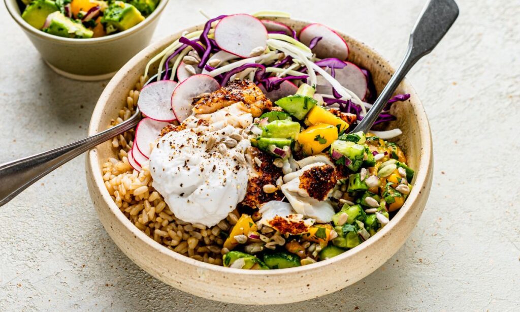 A fertility-friendly burrito bowl in a rustic beige ceramic dish, filled with flaky white fish, brown rice, avocado and mango salsa, sliced radish, shredded red and white cabbage, yoghurt, and sunflower seeds, topped with cracked black pepper. The bowl is placed on a light textured surface with a small bowl of avocado salsa in the background.