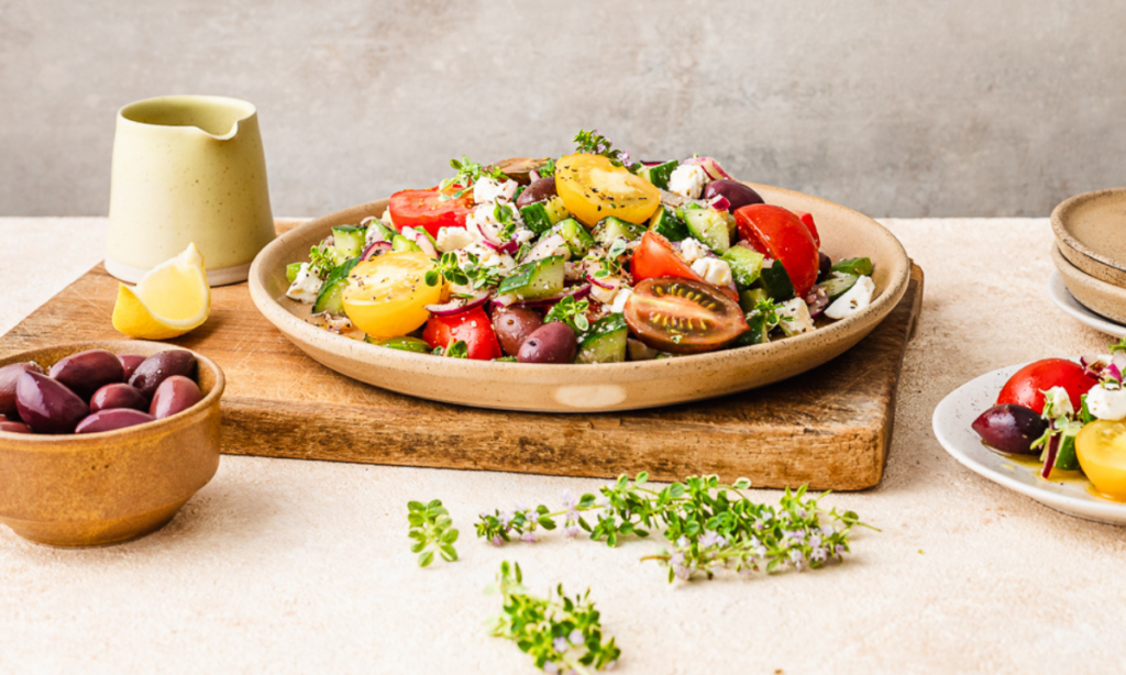 A fertility-friendly Greek salad served on a rustic beige plate, featuring vibrant cherry tomatoes in red, and yellow hues, diced cucumber, Kalamata olives, crumbled feta, and fresh herbs. The plate is set on a wooden board with a bowl of olives, lemon wedge, and scattered fresh herbs in the background, creating a bright, Mediterranean-inspired scene.