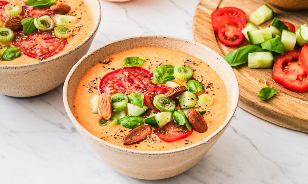 A bowl of fertility-friendly Mediterranean gazpacho, topped with sliced fresh tomatoes, diced cucumber, spring onions, whole almonds, and basil leaves. The vibrant orange soup is served in a rustic beige bowl, set on a white marble surface with a wooden board of fresh tomatoes, cucumber, and basil in the background.