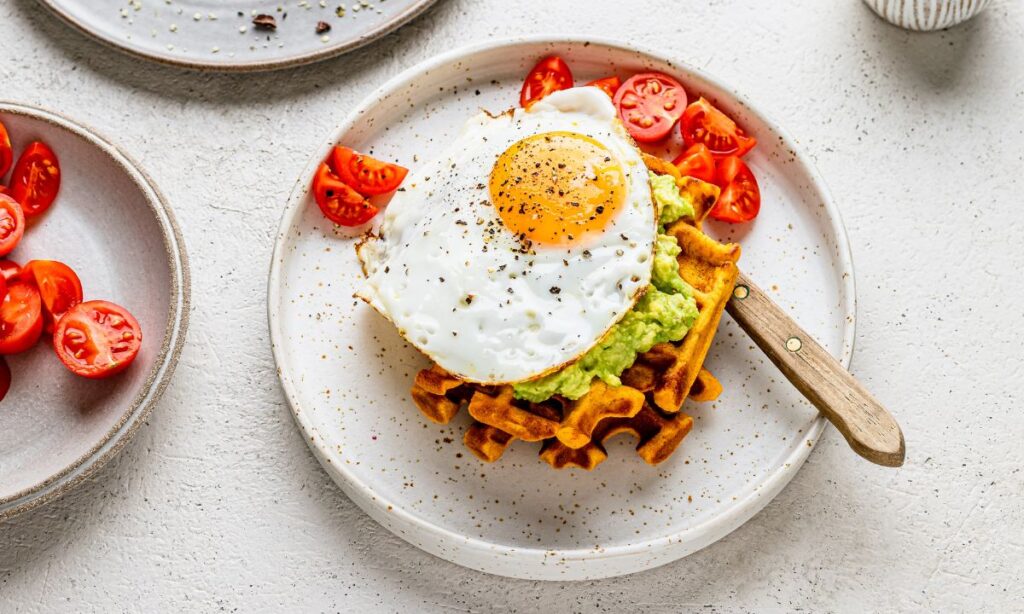 Fried egg on top of sweet potato waffle with smashed avocado, served with cherry tomatoes – a nutrient-dense, fertility-boosting meal rich in protein and healthy fats.