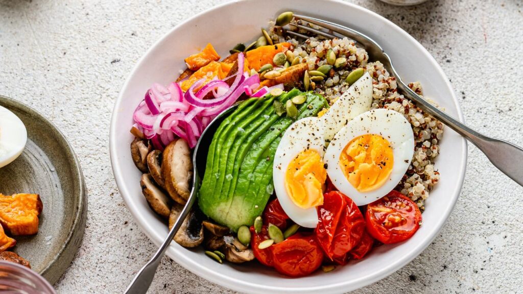 High-protein fertility breakfast bowl featuring quinoa, soft-boiled eggs, avocado, roasted sweet potatoes, sautéed mushrooms, cherry tomatoes, pickled red onions, and pumpkin seeds. A nutrient-dense meal packed with protein, healthy fats, and fibre to support hormone balance and fertility.