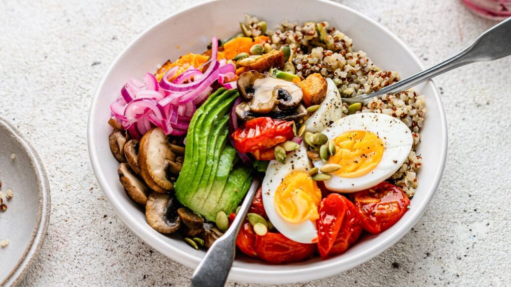 A close-up of a colourful fertility abundance bowl served in a white ceramic dish on a textured light-coloured surface. The bowl contains a mix of nourishing ingredients: fluffy quinoa as a base, topped with sliced creamy avocado, golden soft-boiled eggs, sautéed mushrooms, roasted cherry tomatoes, tangy pickled red onions, and crunchy pumpkin seeds. A silver fork rests in the bowl, ready to take a bite. The vibrant colours and textures of the ingredients create an inviting and nutrient-dense meal designed to support balanced blood sugar and optimise fertility.