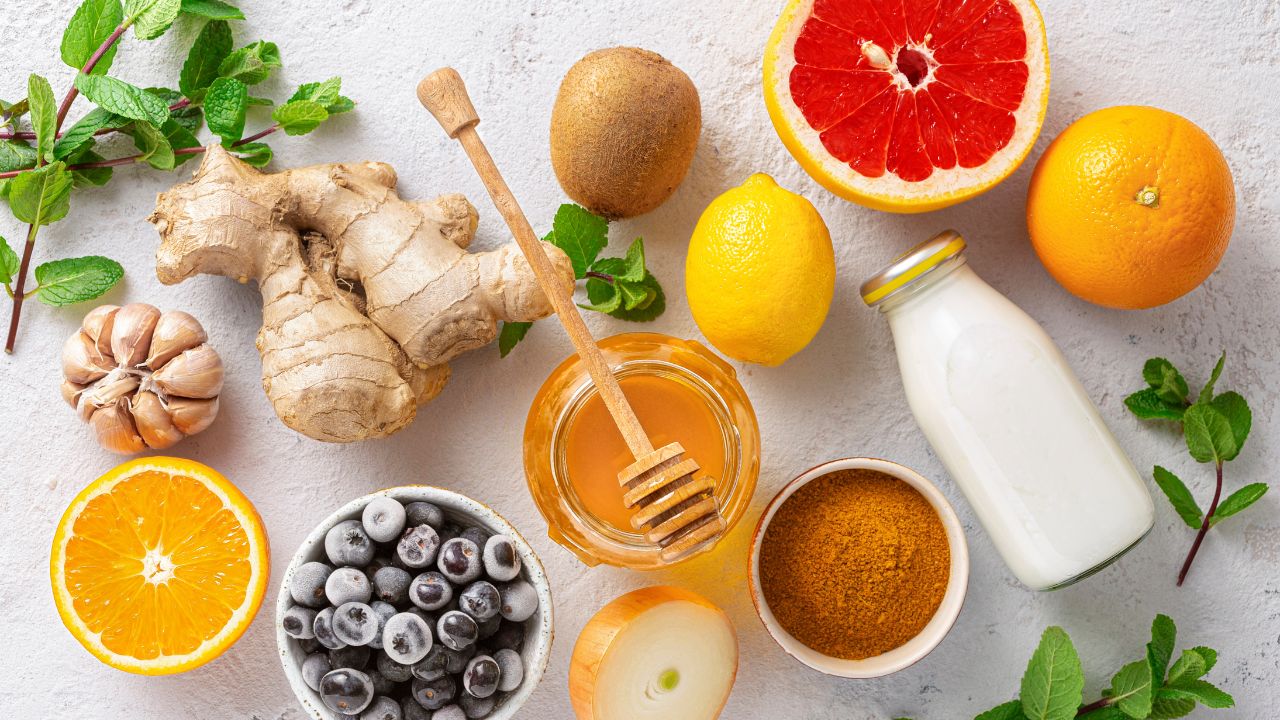 A flat lay of immune-boosting foods arranged on a light textured surface, including fresh ginger, garlic, honey, turmeric, frozen blueberries, lemon, grapefruit, orange, kiwi, onion, mint leaves, and a bottle of probiotic-rich yogurt. These natural ingredients support immune health with their anti-inflammatory and antioxidant properties.