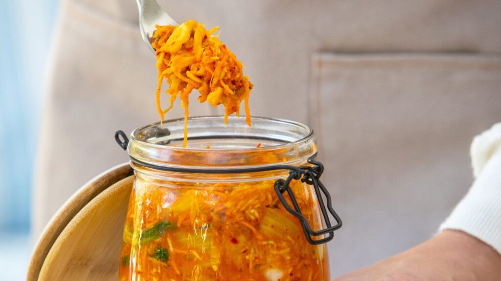 A glass jar of homemade kimchi with a fork lifting a portion, showcasing its vibrant orange color and fermented texture. This probiotic-rich food supports gut health, the immune system and fertility by promoting a healthy microbiome and reducing inflammation.