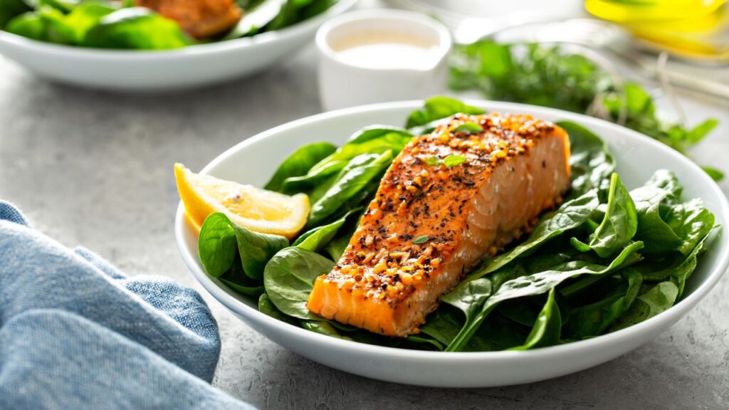 A white bowl of Mediterranean  salmon with a golden crust, seasoned with za'atar - a blend of herbs and spices. The salmon is served on a bed of fresh spinach with a lemon wedge. A creamy dressing and fresh herbs are visible in the background. This nutrient-rich meal is packed with omega-3s, nutrients and antioxidants to support the immune system and fertility.